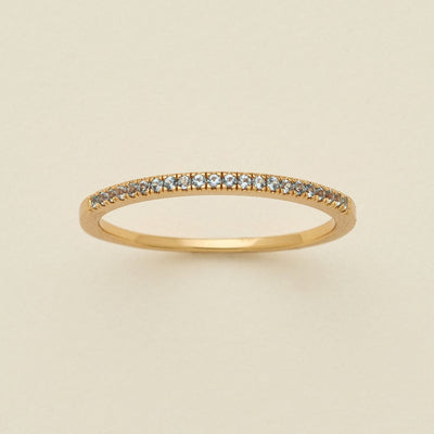 A gold ring featuring a single row of small round-cut light blue-grey gemstones, photographed on a clean, solid, warm off-white background.