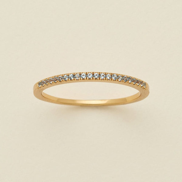 A gold ring featuring a single row of small round-cut light blue-grey gemstones, photographed on a clean, solid, warm off-white background.