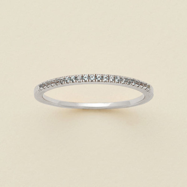 A silver ring featuring a single row of small round-cut light blue-grey gemstones, photographed on a clean, solid, warm off-white background.