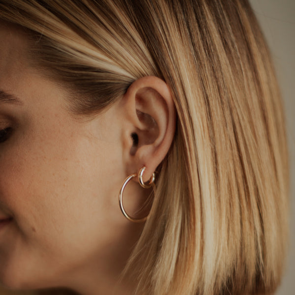 A close-up side profile of a woman with blonde hair wearing a two pairs of delicate gold hoop earrings.