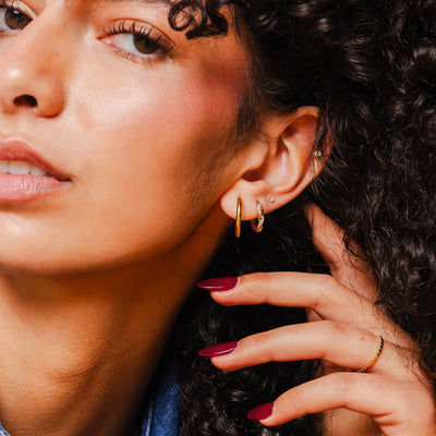 A close-up of a woman's ear wearing a polished gold hoop and a gold braided hoop with small clear accent stones.