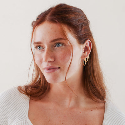 A close-up of a woman in a white knit shirt in front of a solid white background, wearing three different sizes of polished gold hoop earrings.