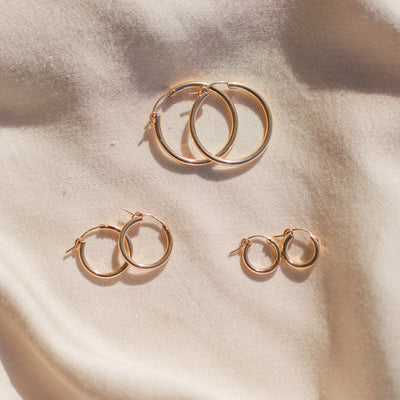 Three pairs of gold hoop earrings arranged on a soft beige fabric background with warm sunlight and shadows.