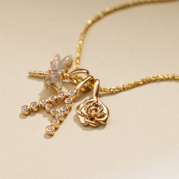 A textured gold chain necklace holding a dragonfly charm with translucent natural gemstones, an initial 'A' charm made up of small round clear stones, and a gold peony flower charm.