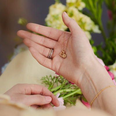 A woman holding a gold pendant necklace with two small flower charms in front of pink, white and green foliage.