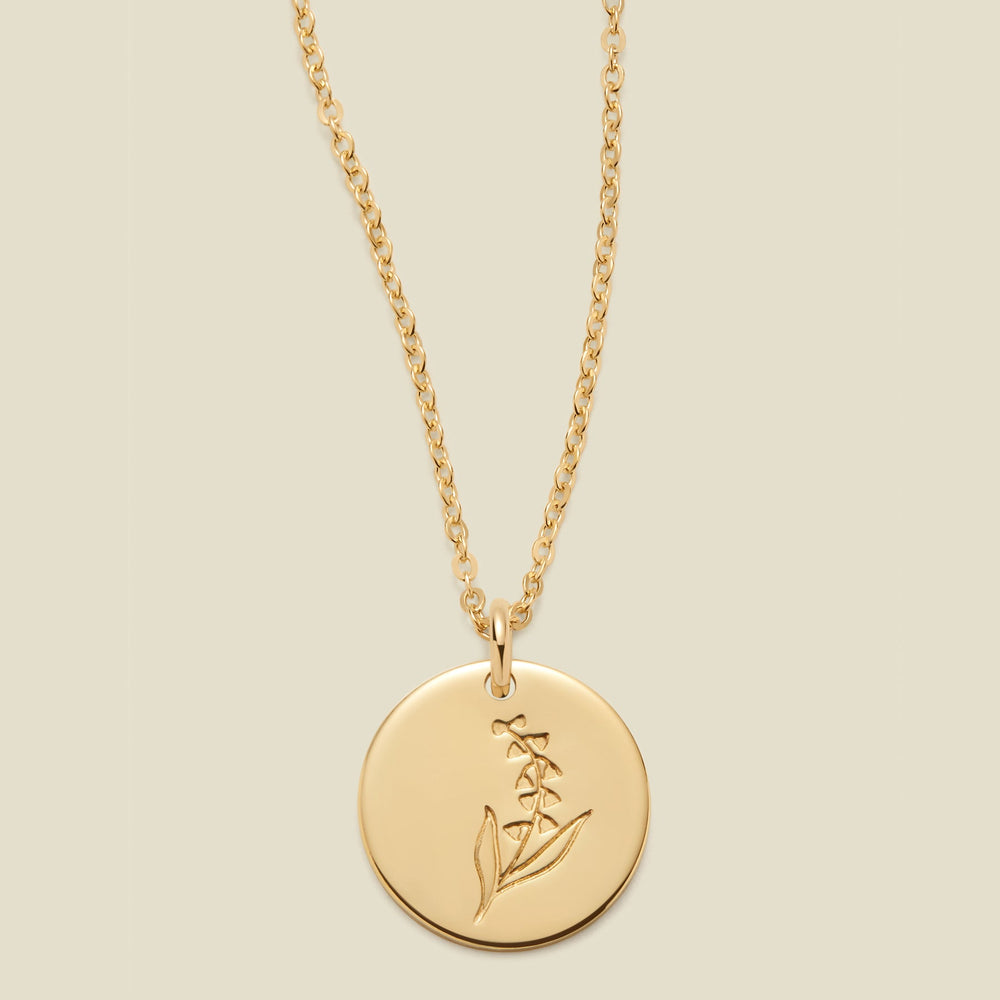 A gold chain necklace with a round gold disc pendant featuring an engraved lily of the valley flower design, displayed against a solid light beige background.