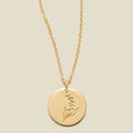 A gold chain necklace with a round gold disc pendant featuring an engraved lily of the valley flower design, displayed against a solid light beige background.