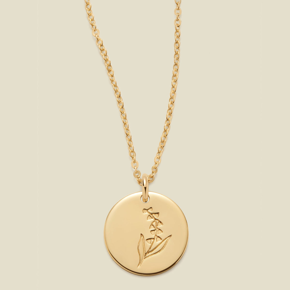 A gold chain necklace with a round gold disc pendant featuring an engraved lily of the valley flower design, displayed against a solid light beige background.