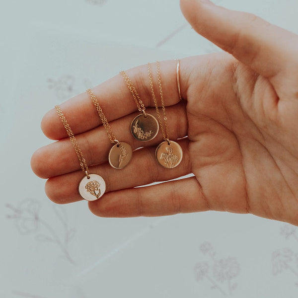A hand holding four gold pendant chain necklaces, each with a flat round disc engrafed with a different floral design.