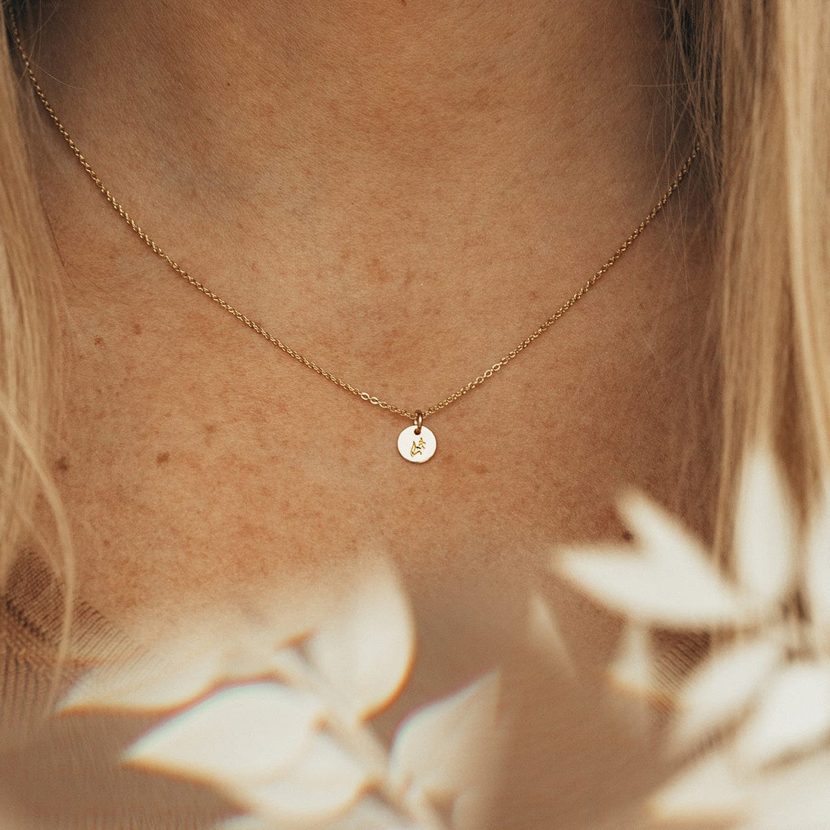 A close-up of a person wearing a dainty gold pendant necklace with a small round flat disc charm. The charm is engraved with a floral motif, and their is beige foliage blurred in the foreground.