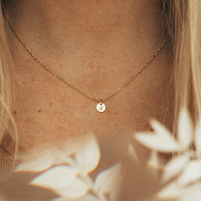 A close-up of a person wearing a dainty gold pendant necklace with a small round flat disc charm. The charm is engraved with a floral motif, and their is beige foliage blurred in the foreground.