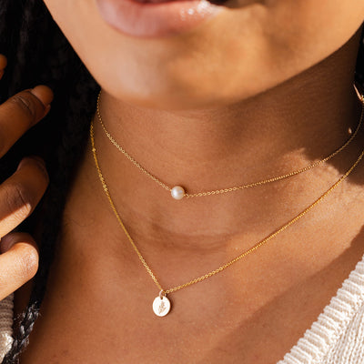 A close-up of a woman wearing a thin gold choker with a single pearl in the center, layered with a delicate gold pendant chain with a flat round disc charm. The charm is engraved with a floral motif.