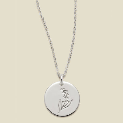 A silver chain necklace with a round silver disc pendant featuring an engraved lily of the valley flower design, displayed against a solid light beige background.