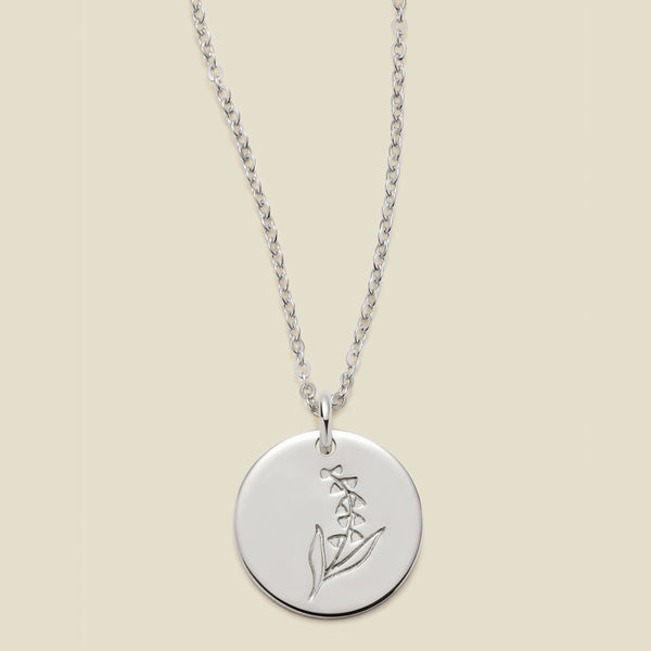 A silver chain necklace with a round silver disc pendant featuring an engraved lily of the valley flower design, displayed against a solid light beige background.