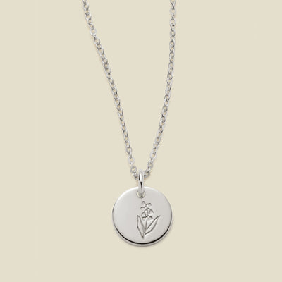 A silver chain necklace with a round silver disc pendant featuring an engraved lily of the valley flower design, displayed against a solid light beige background.