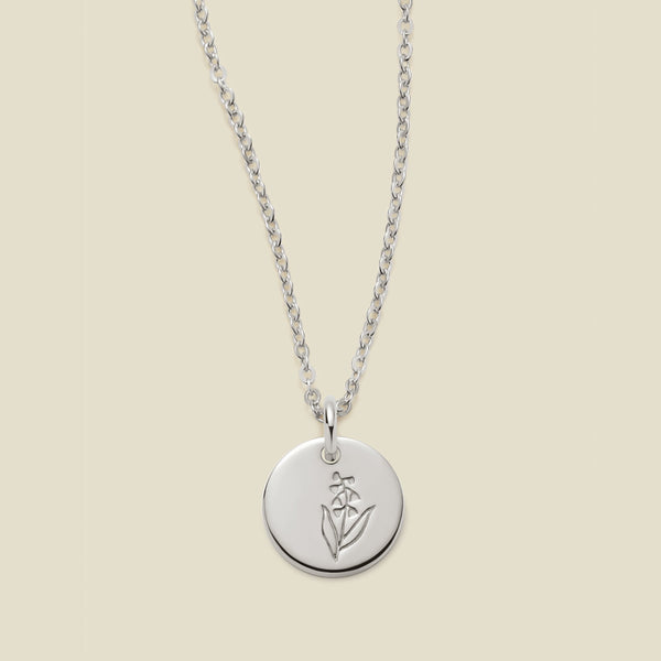 A silver chain necklace with a round silver disc pendant featuring an engraved lily of the valley flower design, displayed against a solid light beige background.