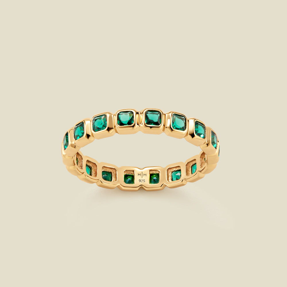 A gold eternity ring featuring a continuous row of square cut dark green gemstones, shown on a solid light beige background.