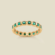 A gold eternity ring featuring a continuous row of square cut dark green gemstones, shown on a solid light beige background.