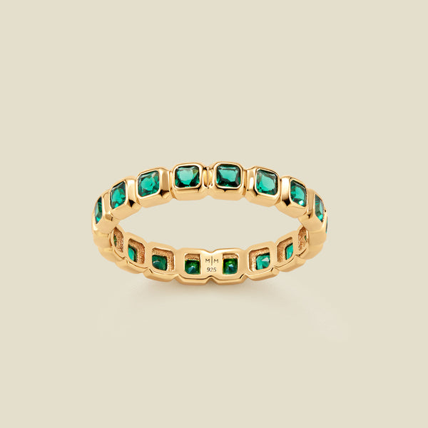 A gold eternity ring featuring a continuous row of square cut dark green gemstones, shown on a solid light beige background.