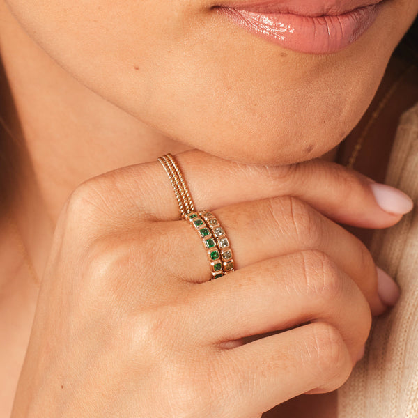 A close-up of a person holding their hand to their chin, wearing three thin stacked gold braided rings and two stacked gold eternity rings, each with either dark green or clear square cut gemstones in a single continuous row.