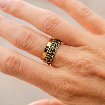 A close-up of a finger wearing four rings stacked: a thick polished gold band, two thin stacked gold rings with small round dark green gemstones in a single row on the front, and a thicker gold eternity ring with dark green square cut gemstones in a continuous row.