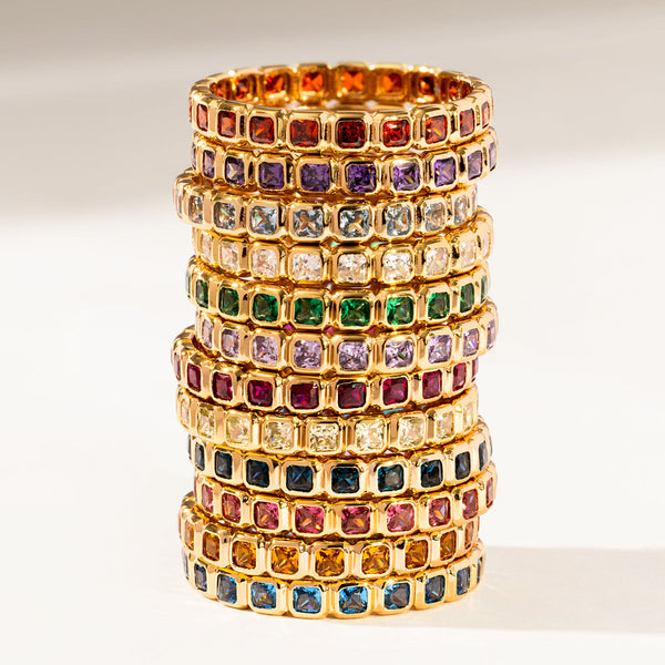 Twelve stacked gold eternity rings, each with a different color of square cut gemstones in a single continuous row, shown on a warm white background.