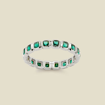 A silver eternity ring featuring a continuous row of square cut dark green gemstones, shown on a solid light beige background.