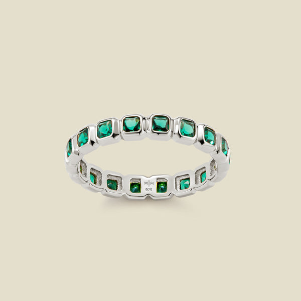 A silver eternity ring featuring a continuous row of square cut dark green gemstones, shown on a solid light beige background.