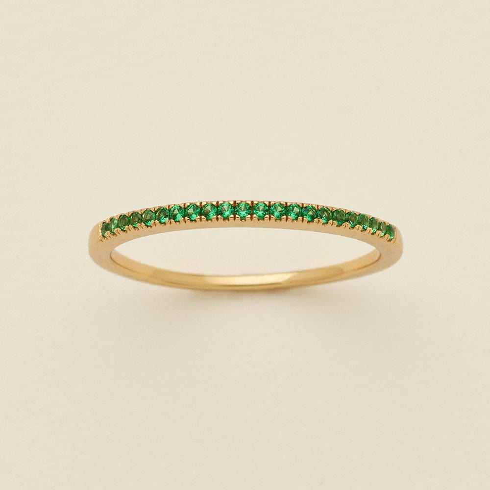 A gold ring featuring a single row of small round-cut green gemstones, photographed on a clean, solid, warm off-white background.