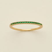 A gold ring featuring a single row of small round-cut green gemstones, photographed on a clean, solid, warm off-white background.