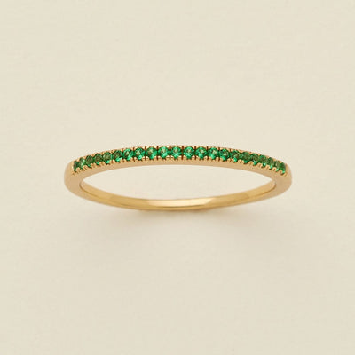 A gold ring featuring a single row of small round-cut green gemstones, photographed on a clean, solid, warm off-white background.