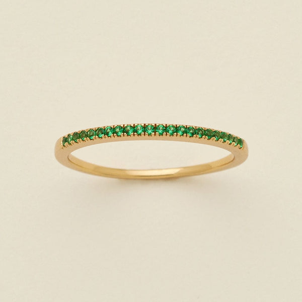 A gold ring featuring a single row of small round-cut green gemstones, photographed on a clean, solid, warm off-white background.