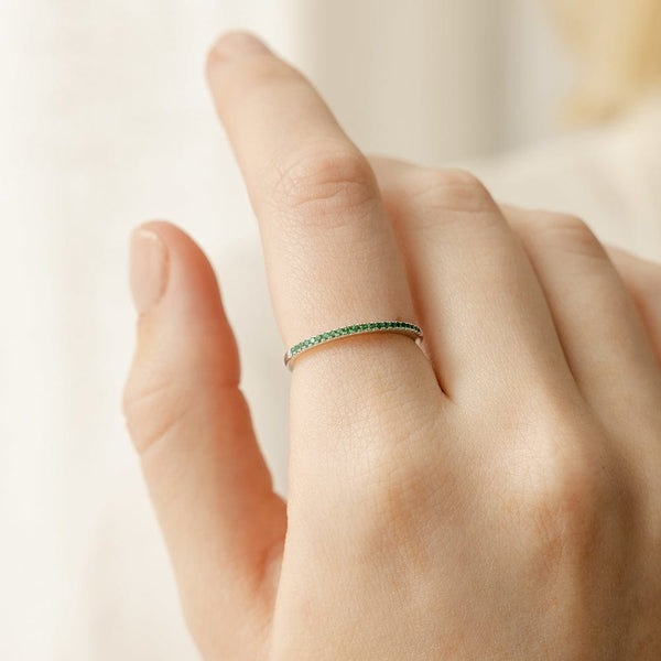 A close-up of a hand with a thin silver ring, featuring a single row of small round-cut green gemstones, set against a light beige blurred background.