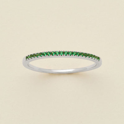 A silver ring featuring a single row of small round-cut green gemstones, photographed on a clean, solid, warm off-white background.