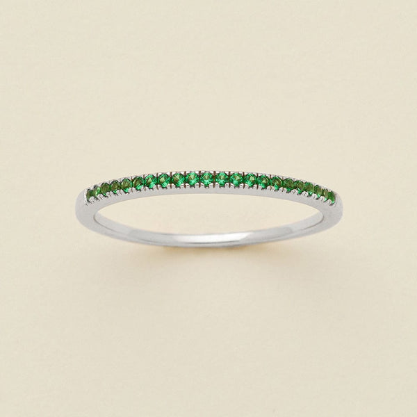A silver ring featuring a single row of small round-cut green gemstones, photographed on a clean, solid, warm off-white background.