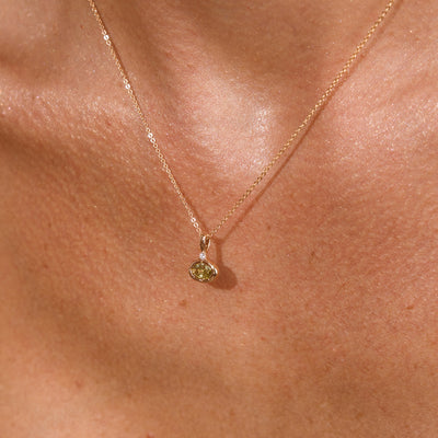 A close up lifestyle photo of a person's chest wearing a dainty gold chain pendant necklace with a gold oval charm featuring an oval bezel-set light green gemstone.