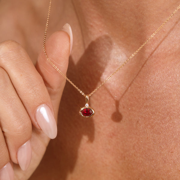 A close up lifestyle photo of a person's neck wearing a dainty gold chain necklace with a gold oval charm featuring a red oval gemstone, being held by a hand to showcase the pendant.
