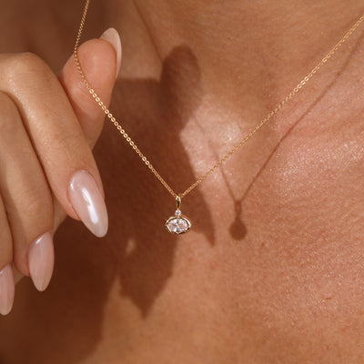 A close up lifestyle photo of a person's neck wearing a dainty gold chain necklace with a gold oval charm featuring a clear oval gemstone, being held by a hand to showcase the pendant.