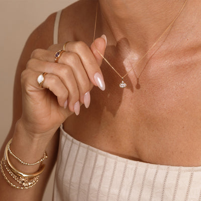 A close up lifestyle photo of a person's neck wearing a dainty gold chain necklace with a gold oval charm featuring a clear oval gemstone, being held by a hand with multiple gold rings and bracelets.