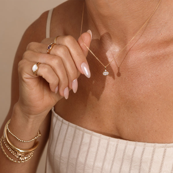 A close up lifestyle photo of a person's neck wearing a dainty gold chain necklace with a gold oval charm featuring a clear oval gemstone, being held by a hand with multiple gold rings and bracelets.