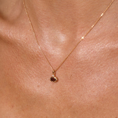 A close up lifestyle photo of a person's neck wearing a dainty gold chain necklace with a gold oval charm featuring a red oval gemstone.