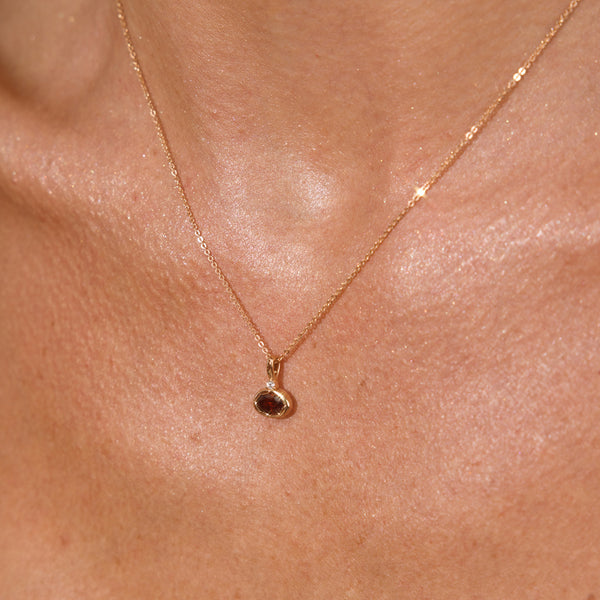 A close up lifestyle photo of a person's neck wearing a dainty gold chain necklace with a gold oval charm featuring a red oval gemstone.