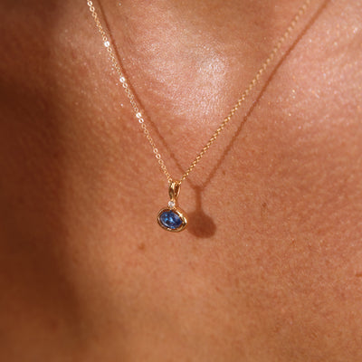 A close up lifestyle photo of a person's chest wearing a dainty gold chain pendant necklace with a gold oval charm featuring an oval bezel-set blue gemstone.