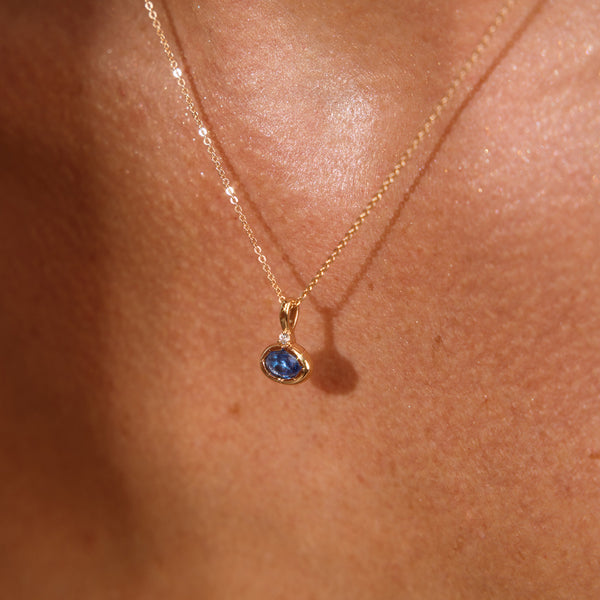 A close up lifestyle photo of a person's chest wearing a dainty gold chain pendant necklace with a gold oval charm featuring an oval bezel-set blue gemstone.