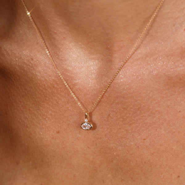 A close up lifestyle photo of a person's chest wearing a dainty gold chain pendant necklace with a gold oval charm featuring an oval bezel-set clear gemstone.
