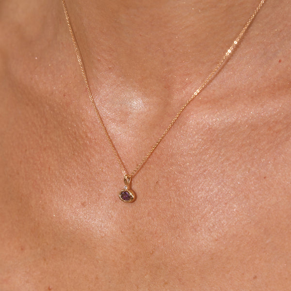 A close-up of a model wearing a dainty gold necklace with a gold oval charm set with a purple gemstone.
