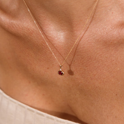 A close up lifestyle photo of a person's chest wearing a dainty gold chain pendant necklace with a gold oval charm featuring an oval bezel-set red gemstone.