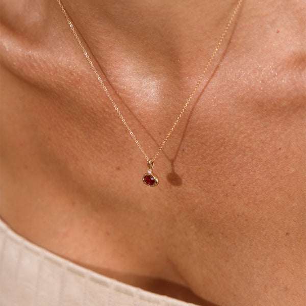 A close up lifestyle photo of a person's chest wearing a dainty gold chain pendant necklace with a gold oval charm featuring an oval bezel-set red gemstone.