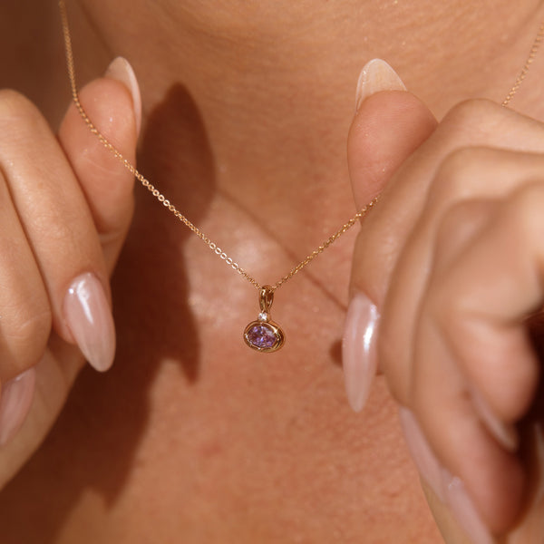 A close up lifestyle photo of a person's neck wearing a dainty gold chain necklace with a gold oval charm featuring a light purple oval gemstone, being held by two hands to showcase the pendant.