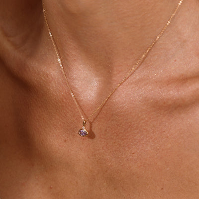 A close up lifestyle photo of a person's chest wearing a dainty gold chain pendant necklace with a gold oval charm featuring an oval bezel-set light purple gemstone.